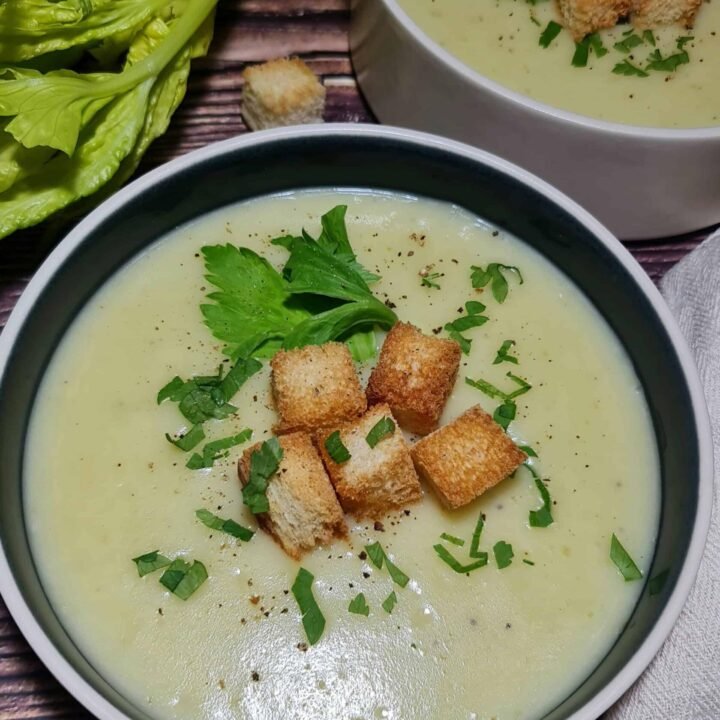 Two rustic bowls of creamy, light green potato celery soup, garnished with golden-brown croutons and fresh parsley, set on a dark wood table.