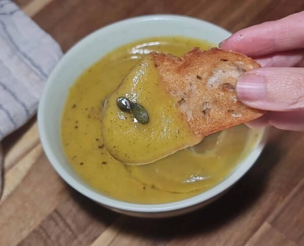 Toasted bread dipped into creamy pumpkin zucchini soup no cream with pumpkin seeds.