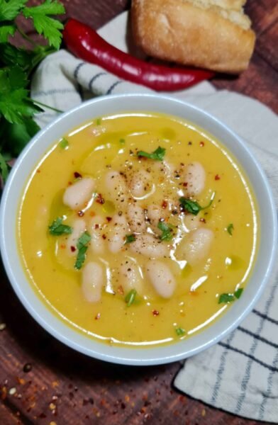 Close-up of a creamy yellow cauliflower soup topped with white beans, parsley, chili flakes, black pepper, and a drizzle of olive oil.