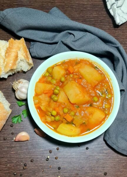 A rustic, overhead shot of a bowl of chunky vegan potato and peas soup, served with crusty bread, garlic, and black peppercorns on a dark wooden surface.