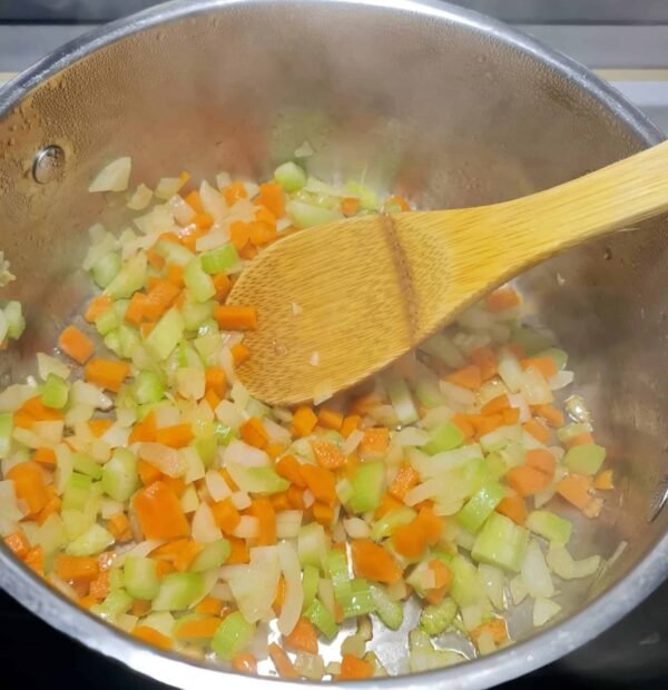 Diced onions, celery, and carrots being sautéed in a silver pot with a wooden spoon, forming the flavor base for the potato and peas soup.
