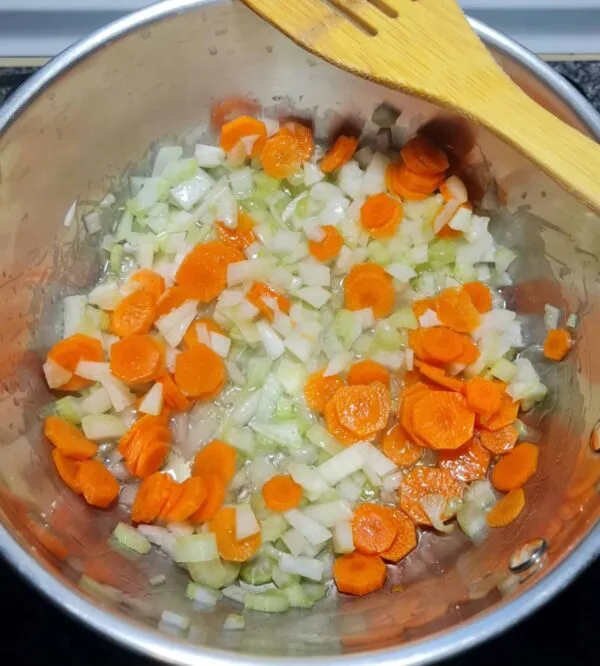 Diced onions, sliced carrots, and celery being sautéed in a silver pot with a wooden spoon, forming the flavorful aromatic base for the spinach noodle soup.