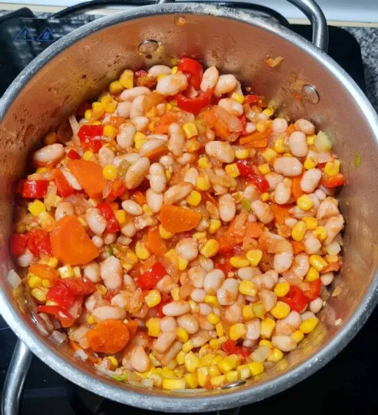 White beans, corn, carrots, and red peppers cooking in a pot on the stovetop, a critical step for preparing the vegan bean and corn soup.