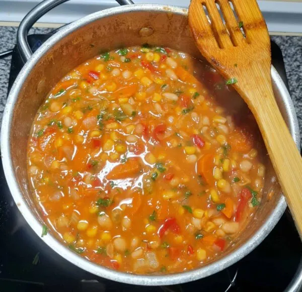 The vegan bean and corn soup is simmering in a silver pot on the stove, with a wooden spoon resting on the side, demonstrating the final cooking stage.