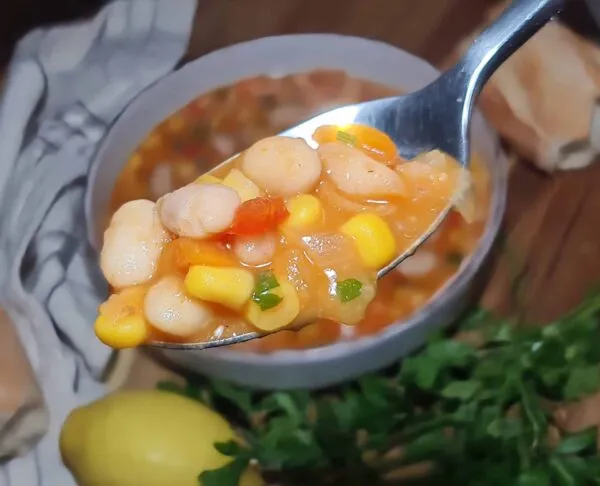 Extreme close-up of a silver spoon lifting a portion of hot, steaming vegan bean and corn soup, showing the texture of the beans and corn.