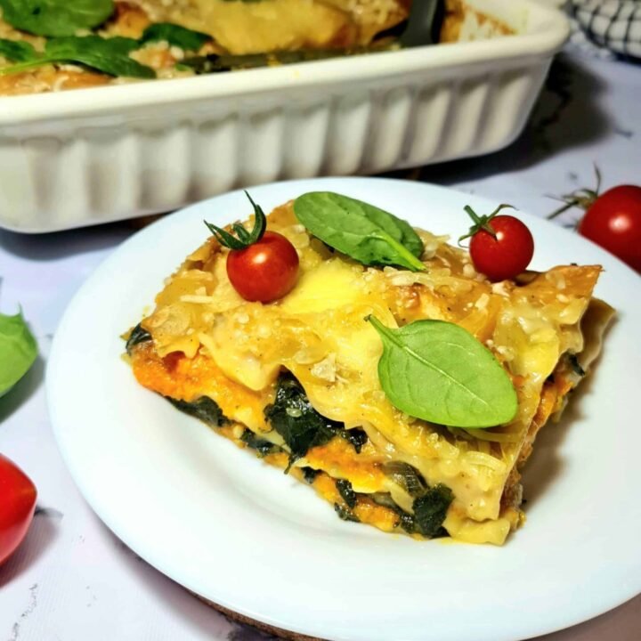 Slice of vegan pumpkin spinach lasagna on a white plate with cherry tomatoes and spinach leaves.