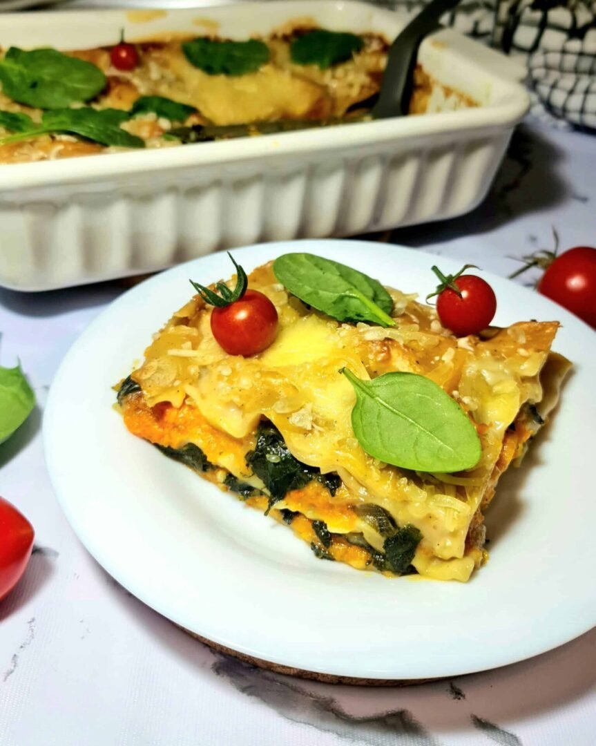 Slice of vegan pumpkin spinach lasagna on a white plate with cherry tomatoes and spinach leaves.
