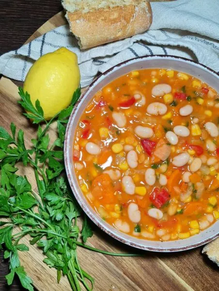A close-up shot of a bowl of colorful vegan bean and corn soup, garnished with fresh parsley and served with crusty bread.