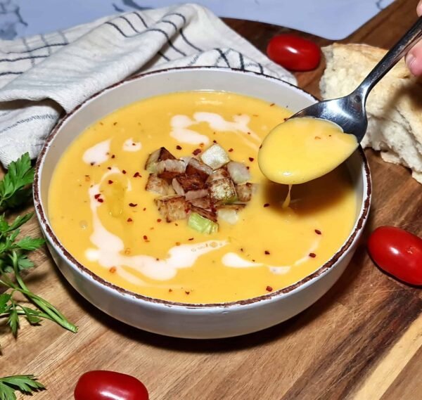 A close-up bowl of silky winter kohlrabi soup, topped with golden crispy kohlrabi cubes and a swirl of coconut cream. A hand lifts a spoonful to show its smooth, velvety texture.