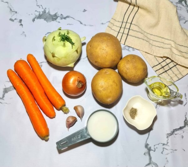 Overhead shot of the ingredients for winter kohlrabi soup, including kohlrabi, carrots, potatoes, garlic, onion, olive oil, coconut milk, and spices arranged on a marble surface.